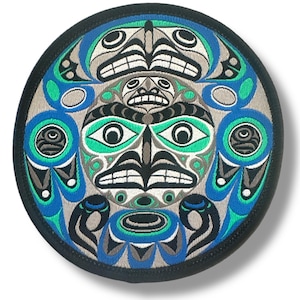 Puede incluir: Parche bordado redondo con un diseño nativo americano estilizado. El parche tiene un borde negro y un diseño central en tonos de azul, verde, gris y negro. El diseño incluye formas y caras simétricas.