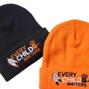 May include: A black knitted beanie and an orange knitted beanie, each featuring an embroidered design with the text "EVERY CHILD MATTERS," a graphic of two feathers, and a handprint icon. The black beanie has orange text, while the orange beanie has black text.