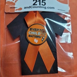 May include: A circular orange lapel pin featuring the text EVERY CHILD MATTERS, a dreamcatcher graphic, and two small white footprints. The pin is attached to a black t-shirt shaped card with an orange ribbon, packaged in a clear plastic sleeve.