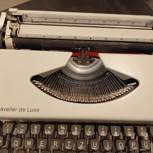 Puede incluir: Una máquina de escribir vintage AEG Olympia Traveller de Luxe. La máquina de escribir tiene un cuerpo color crema con teclas negras y un carro plateado. El texto "Traveller de Luxe" está impreso en la parte delantera. Las teclas están dispuestas en QWERTY.