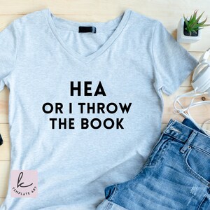 May include: A light gray V-neck t-shirt with the text "HEA OR I THROW THE BOOK" in black. The shirt is paired with denim shorts. A small potted plant and headphones are visible in the background.