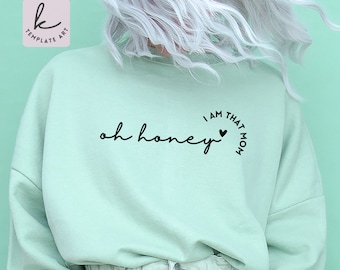 Puede incluir: Una sudadera de color verde claro con el texto "oh honey" y un círculo con el texto "I am that mom" y un corazón.