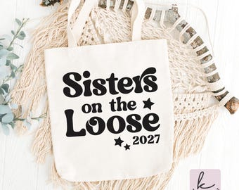 May include: A cream-colored canvas tote bag with two top handles. The front of the bag features black text that reads Sisters on the Loose 2027, accompanied by two small black star icons positioned near the text and the year.