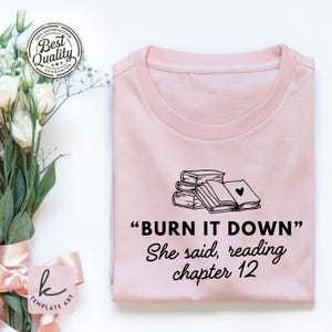 Può includere: T-shirt rosa chiaro con la scritta "BURN IT DOWN" e "She said, reading chapter 12" in nero. La maglietta presenta una grafica di libri impilati con un cuore su un libro aperto. Un adesivo "Best Quality" è in alto a sinistra.