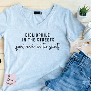 May include: A light gray V-neck t-shirt with the text "BIBLIOPHILE IN THE STREETS feral reader in the sheets" in black. The shirt is paired with denim shorts. A small succulent plant and headphones are visible in the background.