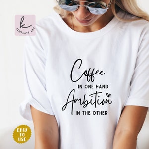 以下が含まれることがあります： 黒い文字で「Coffee in one hand Ambition in the other」と書かれた白いTシャツ。小さなハートのマークが付いている。