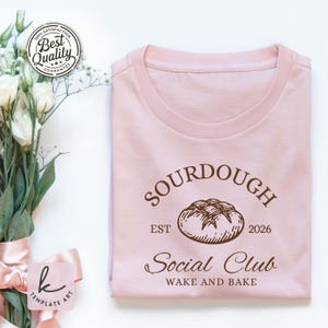 May include: A light pink t-shirt with the words "SOURDOUGH Social Club" and "WAKE AND BAKE" printed in brown. A brown illustration of a loaf of bread is centered above the words. The text "EST 2026" is also printed on the shirt.