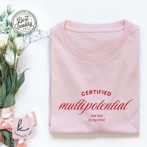 May include: A light pink t-shirt with the words "CERTIFIED multipotential ...but lost in my mind" printed in red. The shirt is folded neatly. A bouquet of white flowers and a pink tag are in the background.