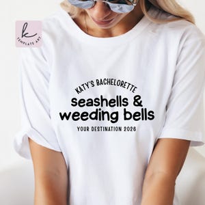 May include: White t-shirt with the text "Katy's Bachelorette seashells & weeding bells YOUR DESTINATION 2026" in black. The shirt is a crew neck and has rolled-up sleeves. The shirt is being worn by a person.
