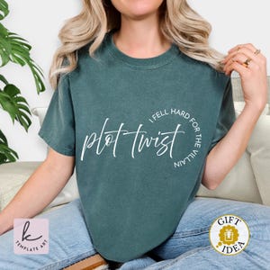 Op de afbeelding: Een tealkleurig t-shirt met de tekst "plot twist" in een cursief lettertype. De zin "I fell hard for the villain" is in een cirkel rond de hoofdtekst geschreven. Het shirt is effen van kleur en heeft korte mouwen.