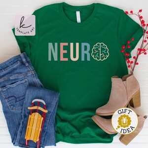 Pode incluir: Camiseta verde com a palavra "NEURO" em letras coloridas e um gr&aacute;fico de c&eacute;rebro. A camiseta est&aacute; sobre uma superf&iacute;cie branca com jeans azul, um tren&oacute; de madeira e botas marrons.