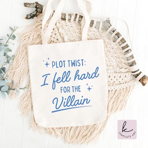May include: A cream-colored tote bag with the text "PLOT TWIST: I fell hard FOR THE Villain" in blue script. The bag has two handles and is made of a canvas-like material. The design includes star accents.