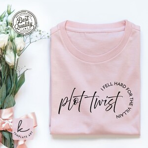 Op de afbeelding: Lichtroze T-shirt met de woorden "plot twist" in een cursief lettertype. De zin "I fell hard for the villain" staat in een cirkel rond de rechterkant van de tekst. Het shirt is netjes opgevouwen.