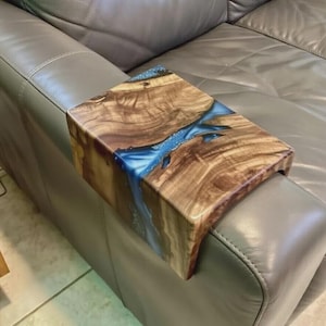 May include: A rectangular wooden sofa arm tray featuring a natural wood grain finish with a central river-like inlay of translucent blue resin. The tray is designed to drape over the arm of a couch, providing a flat surface for items.