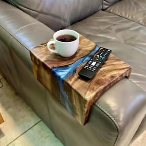 May include: A wooden sofa arm tray with a blue resin river design. The tray holds a white mug of coffee and a black remote control. The tray is placed on a gray leather sofa arm.