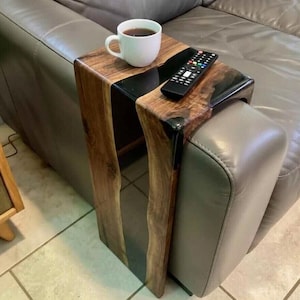 May include: A wooden side table with a live edge design and black resin inlay. A white mug with coffee and a remote control rest on the table, which is placed next to a gray leather sofa.