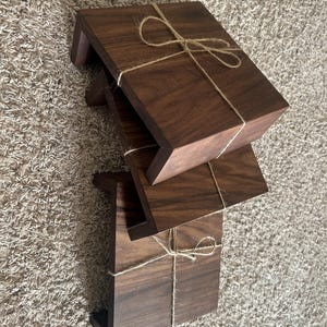 May include: Three stacked, dark brown wooden risers tied together with twine. Each riser has a rectangular top and a lower support, creating a small step. The wood grain is visible, and the twine is tied in a bow.