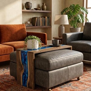 May include: A rectangular wooden side table featuring a central river-like inlay of vibrant blue resin. The table is positioned over a gray leather ottoman with wheels, supporting a white ceramic pot with a trailing green plant and a gray mug.