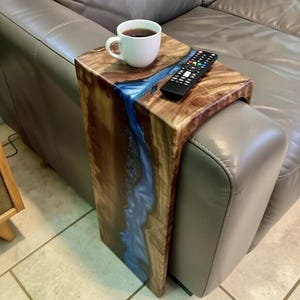May include: A wooden armrest table featuring a natural wood grain finish and a central inlay of translucent blue resin. The table is draped over a gray leather sofa arm and holds a white ceramic coffee mug and a black television remote control.