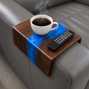 May include: A wooden sofa arm tray with a blue resin river design. A white coffee cup with steam rising sits on the tray, next to a black remote control. The tray is designed to fit over a sofa arm.
