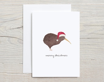 Op de afbeelding: Een kerstkaart met een bruine kiwi-vogel die een rode kerstmanmuts draagt. De kiwi kijkt naar rechts en heeft een lange snavel. De tekst "merry christmas" staat onder de kiwi.