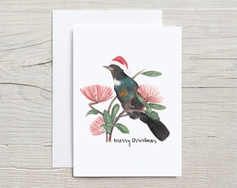 Op de afbeelding: Een kerstkaart met een aquarel-illustratie van een zwart-groene vogel met een rode kerstman-hoed op een tak met roze bloemen. De tekst "merry christmas" staat onder de vogel.