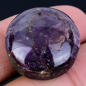 Peut inclure: Une pierre précieuse polie de forme ronde, taillée en cabochon, présentant une palette de couleurs violet foncé et pourpre profond. La pierre possède une surface lisse et réfléchissante avec des inclusions naturelles visibles.