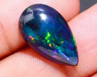 May include: A polished, teardrop-shaped black opal gemstone. The opal displays vibrant internal colors of green, blue, and orange, creating a play-of-color effect. The gemstone is held in a hand, showcasing its size and shape.