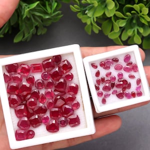 Könnte beinhalten: Zwei weiße quadratische Tabletts gefüllt mit polierten, tiefroten Rubinen in verschiedenen Formen und Größen. Die Rubine werden in einer Hand präsentiert, die ihre leuchtende Farbe und Facetten zeigt. Die Edelsteine sind eine Mischung aus runden, quadratischen und ovalen Schliffen.