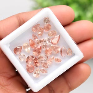 May include: A collection of peach-colored gemstones in various shapes, including teardrops and ovals, are displayed in a small white square container. The gemstones have a translucent quality and are set against a neutral background.