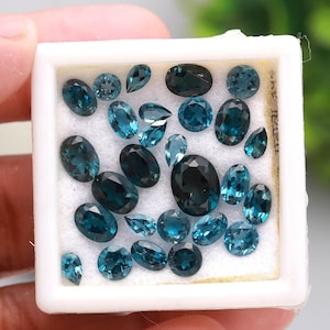 May include: A collection of teal-colored gemstones in various shapes, including oval, pear, and round cuts, displayed in a white box. The gemstones vary in size and shade, with some appearing darker than others. The box is held by a hand.