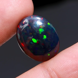 May include: A polished, oval-shaped black opal gemstone. The opal displays vibrant flashes of green, red, and blue, creating a play-of-color effect. The gemstone is held between two fingers, showcasing its size and luster.