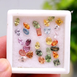 May include: A collection of colorful gemstones in various shapes, including oval, pear, and square cuts, displayed in a white box. The gems are in shades of pink, orange, green, yellow, and blue.