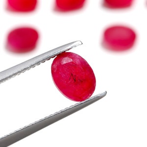 May include: A bright red oval ruby gemstone held in silver tweezers. The ruby is faceted and has a slight shine.