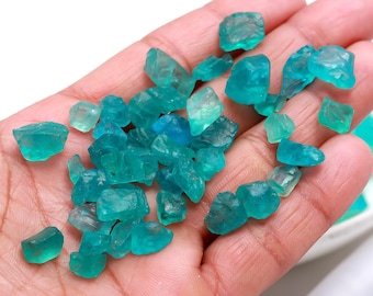 May include: A handful of teal blue apatite crystals, some with a slight green hue, are shown in a person's hand. The crystals are rough and have a natural, unpolished look.