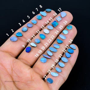 May include: A hand holds 34 opal gemstones, each with a unique shape and size. The opals vary in colour, from pale to deep blue, with some showing iridescent flashes. Each stone is numbered for easy identification.