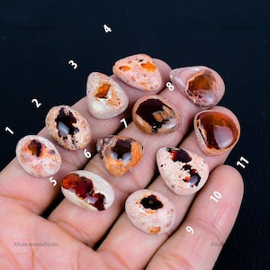 May include: A collection of eleven polished opal gemstones, each with unique patterns and colors. The opals display a mix of creamy white, peach, and deep reddish-brown hues. The gemstones are held in a hand against a black background.