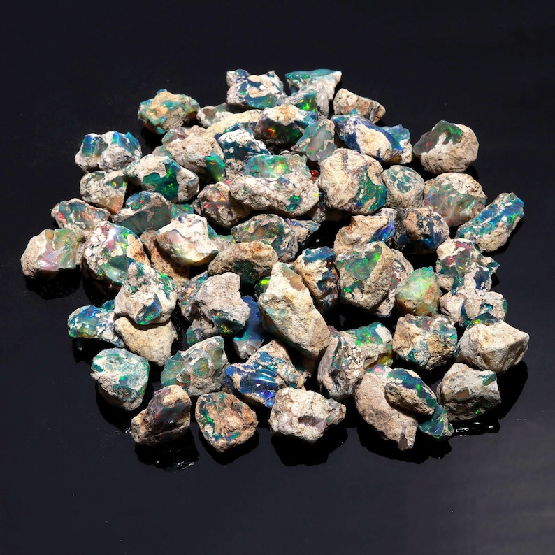 A collection of raw black Ethiopian opal rough stones is scattered on a dark reflective surface.