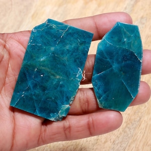 May include: Two pieces of teal blue apatite, one rectangular and one irregularly shaped, with a rough, natural finish. The stones are held in a hand.