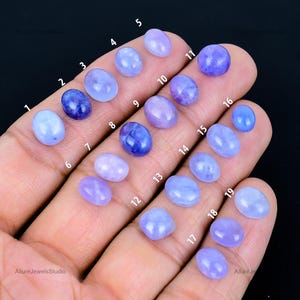 Könnte beinhalten: Eine Sammlung von neunzehn ovalen und runden Edelsteinen in Blau- und Lilatönen, die auf einer Hand präsentiert werden. Die Edelsteine variieren in der Farbintensität, von hellem Periwinkle bis zu tiefem Indigo. Das Bild ist mit "Allure Jewels Studio" beschriftet.
