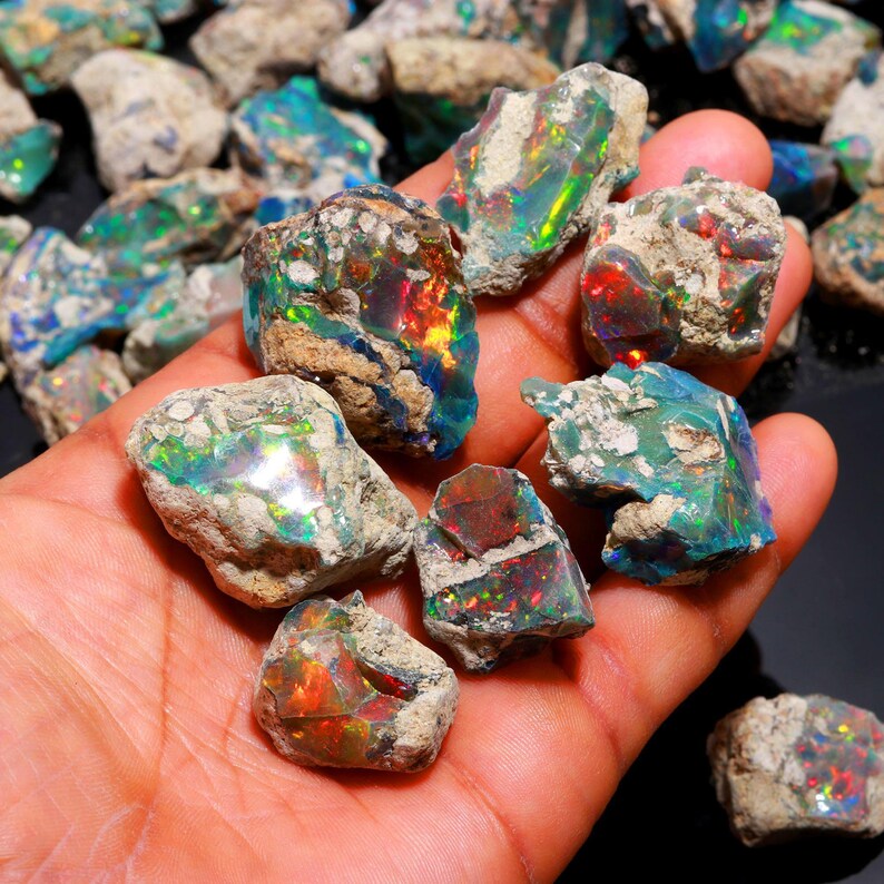 A hand holds several rough black Ethiopian opal stones displaying vibrant flashes of green, red, and blue color play.
