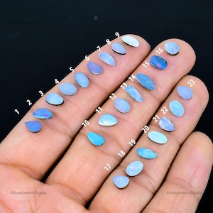 Könnte beinhalten: Eine Sammlung von 23 Opalen in verschiedenen Formen und Größen, die auf einer Hand präsentiert werden. Die Opale reichen in der Farbe von hellblau bis irisierend, wobei einige Farbflecken aufweisen. Jeder Opal ist nummeriert.