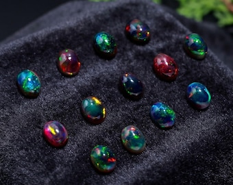 Puede incluir: Una colección de doce ópalos negros de forma ovalada dispuestos en una cuadrícula. Cada piedra preciosa presenta un color base oscuro con vibrantes destellos iridiscentes de luz verde, azul, roja y amarilla reflejados en las superficies pulidas.