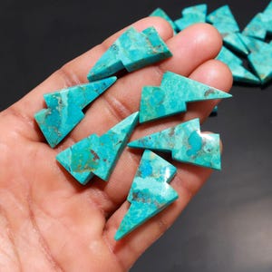 May include: A collection of turquoise lightning bolt shaped cabochons. The cabochons are a vibrant blue-green color with brown and black marbling.