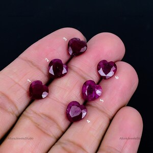 May include: Six heart-shaped, faceted ruby gemstones in varying shades of red and pink are displayed. The gemstones are numbered 1 through 6. The gemstones are held in a hand against a black background.