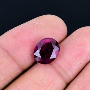 May include: A deep magenta-colored oval gemstone with a faceted surface. The stone features a symmetrical cut with multiple triangular and kite-shaped facets that reflect light, creating a brilliant appearance against the dark background.