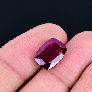 May include: A cushion-cut gemstone with a deep, saturated red color and a faceted surface that reflects light. The stone features a rectangular shape with rounded corners and a polished finish, displaying internal clarity and vibrant red hues.