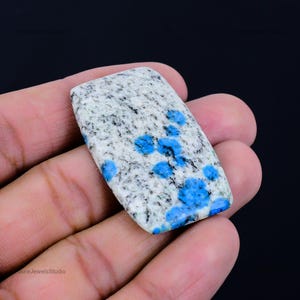 Puede incluir: Un cabujón rectangular de piedra K2 con una base moteada blanca y gris similar al granito, con distintas inclusiones circulares de color azul vibrante dispersas por la superficie. La piedra pulida tiene bordes suaves y redondeados.
