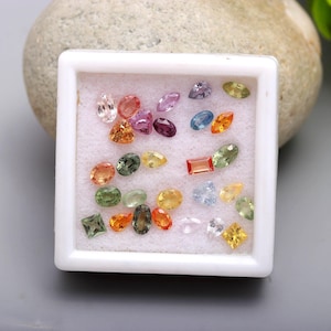 May include: A collection of loose, multi-colored gemstones in various shapes including oval, pear, round, square, and marquise cuts. The stones are arranged on a white foam insert within a square white display box, featuring hues of yellow, orange, green, blue, purple, and clear.