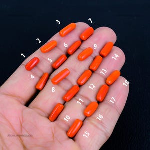May include: Eighteen orange-red coral beads are shown in a hand, each numbered. The beads are oblong and polished. The background is dark, accentuating the coral's colour. The beads are likely measured in centimetres.
